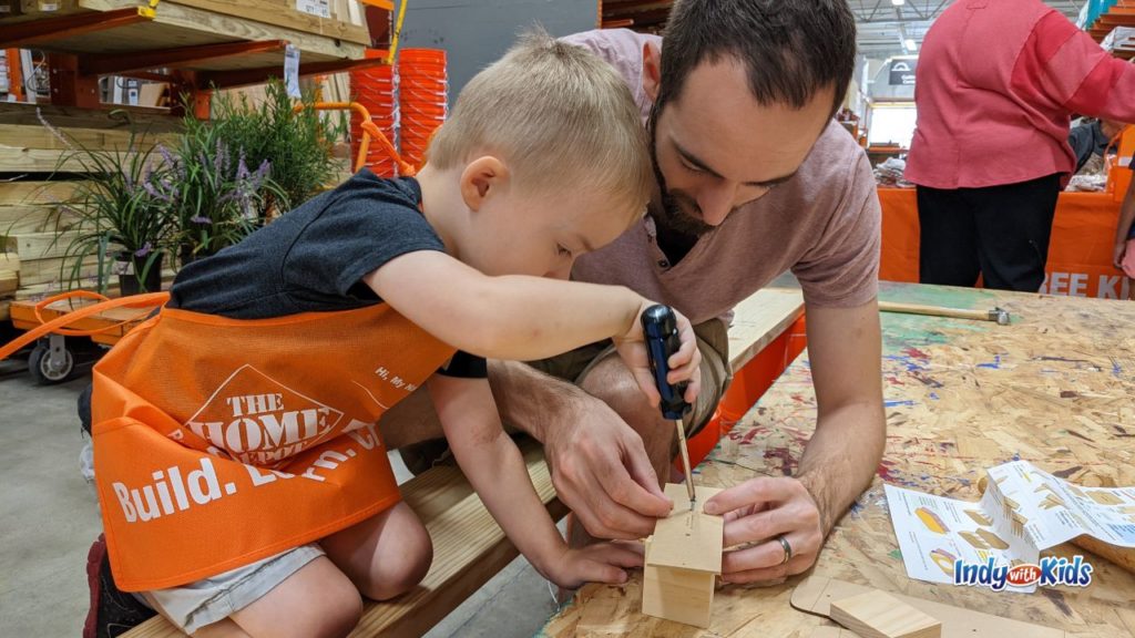 a father holds together pieces of wood as his son, wearing a home depot orange apron, screws in a nail at a home depot kids workshop