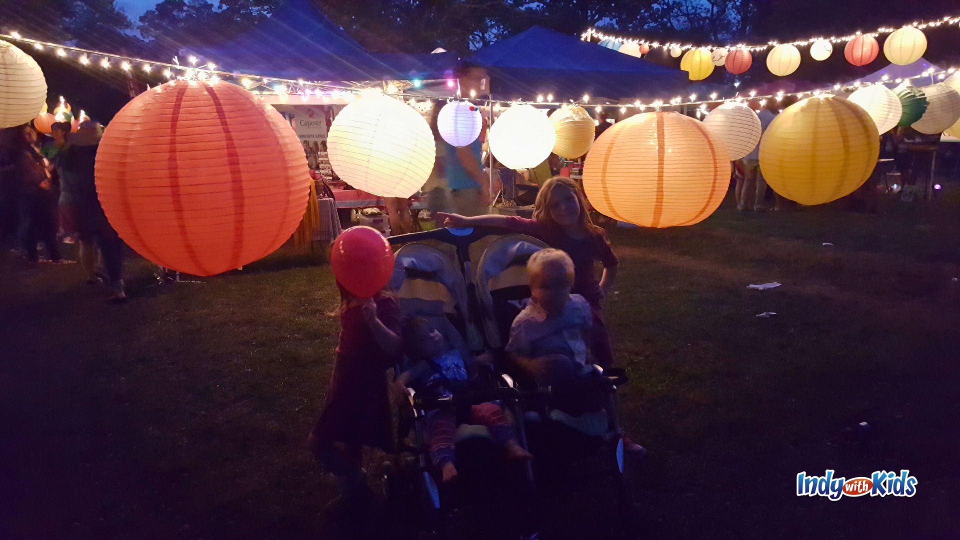 Young children stand beside a double stroller beneath large glowing orange, white, and yellow paper lanterns at the Feast of Lanterns festival in Indianapolis.