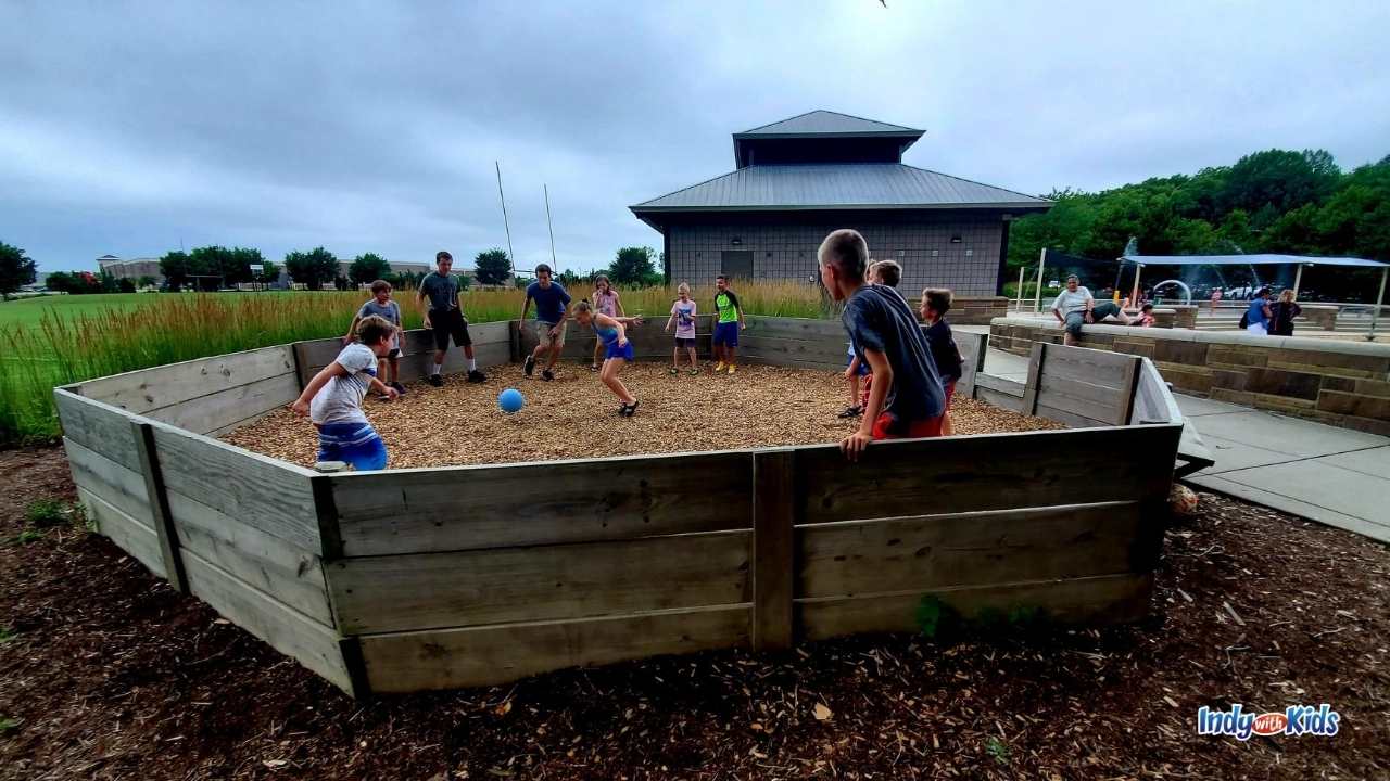 about 12 school-aged children play in an octogonal gaga ball pit made of wood and filled with wood chips at dillon park