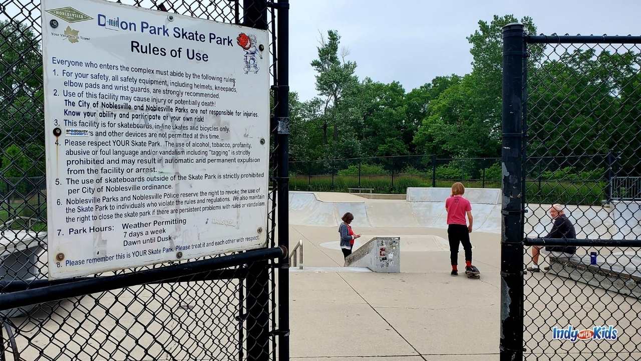 the rules to the skate park are posted on a metal sign hanging on the black fence enclosing the skate park. 3 individuals are inside the skate park. 