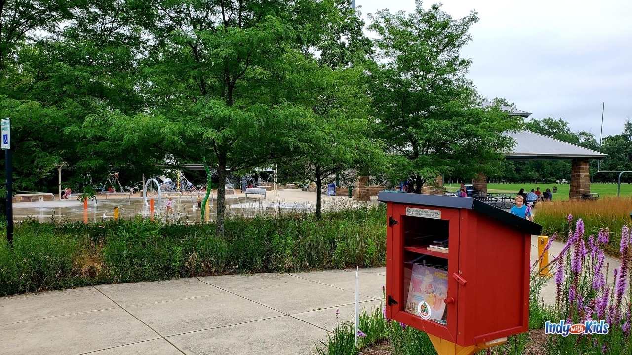 a little wooden red library holds books for exchange at dillon park. the splash pad and park shelter are in the background of the photo.