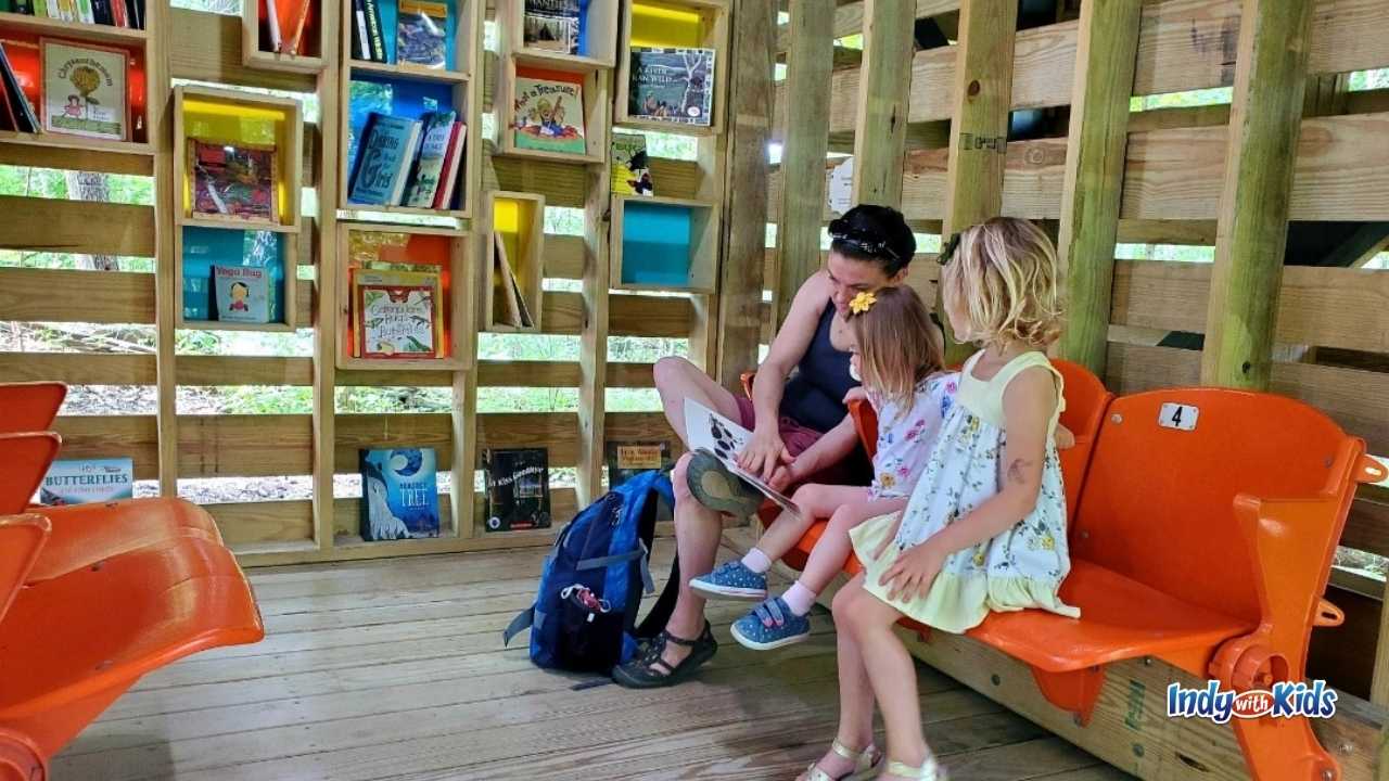 Outdoor Activities for Kids: A woman reads a book to two children while seated on orange folding chairs inside the Treetop Outpost at Conner Prairie.
