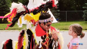 A dancer wearing colorful feather regalia smiles while speaking with a young child at an outdoor powwow.
