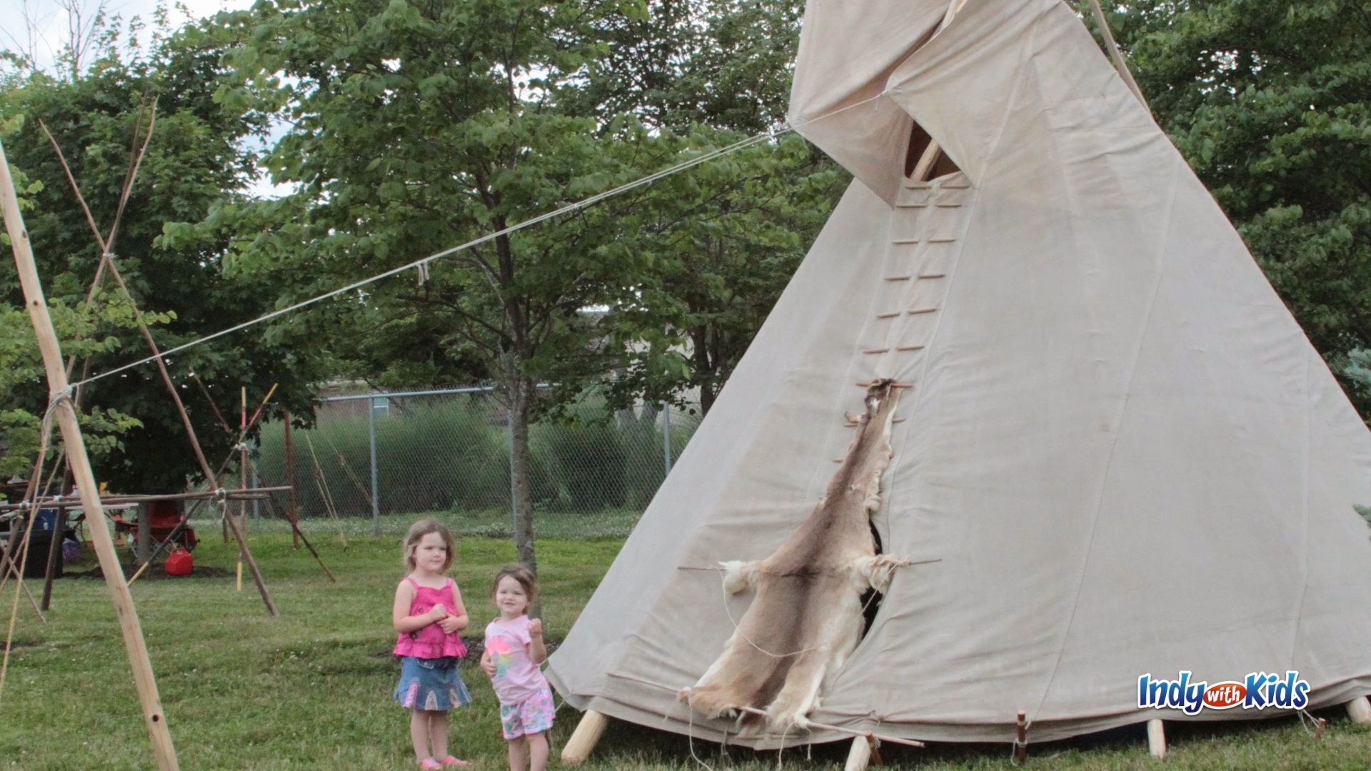 Two young children stand outside a large canvas tipi in a grassy encampment area.