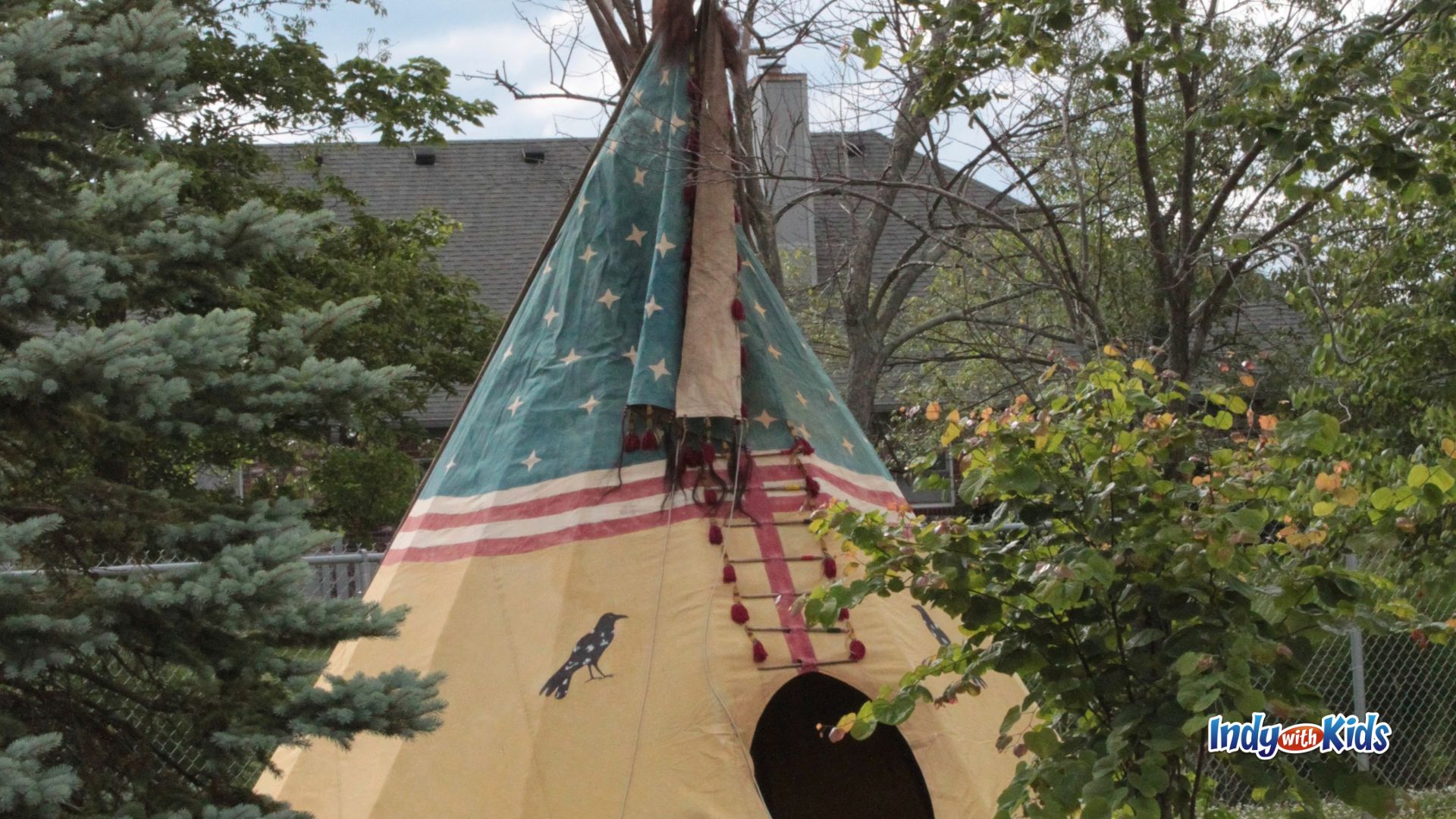 A painted tipi with stars, stripes, and bird designs stands among trees at an outdoor encampment.