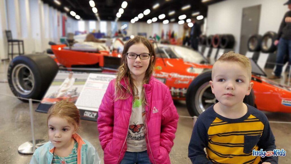 Three children stand in front of a historic Indianapolis 500 race car inside of the Indianapolis Motor Speedway Museum.