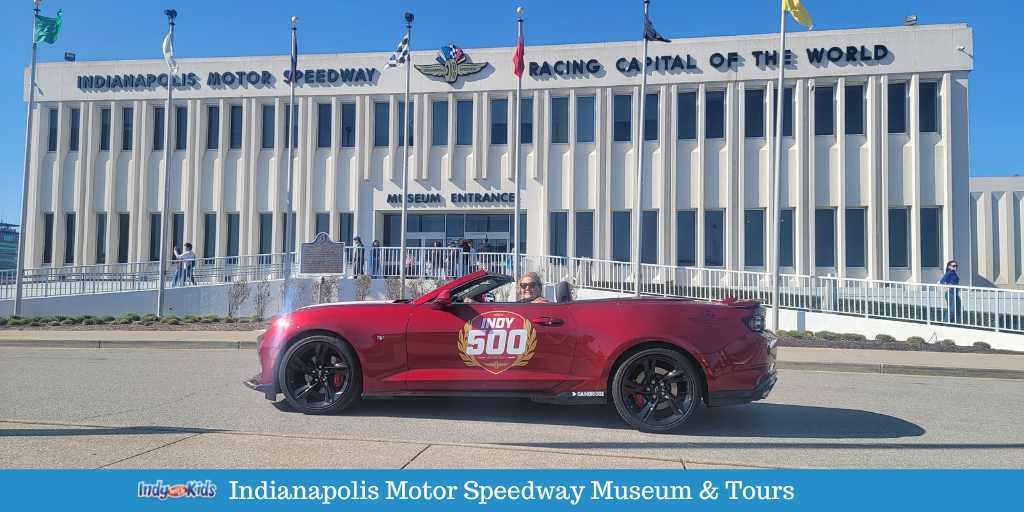 A red indianapolis 500 pace car is parked in front of the Indianapolis motor speedway museum.