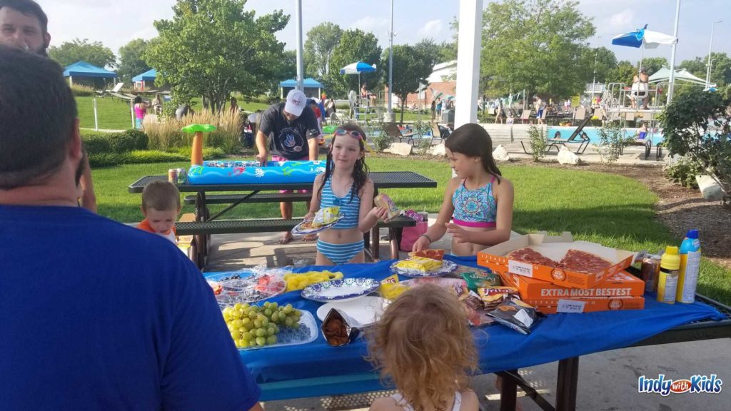 Carmel Water Park: a family is enjoying grapes, pizza, and chips around a picnic table with a blue tablecloth on it for a Birthday party at The Waterpark