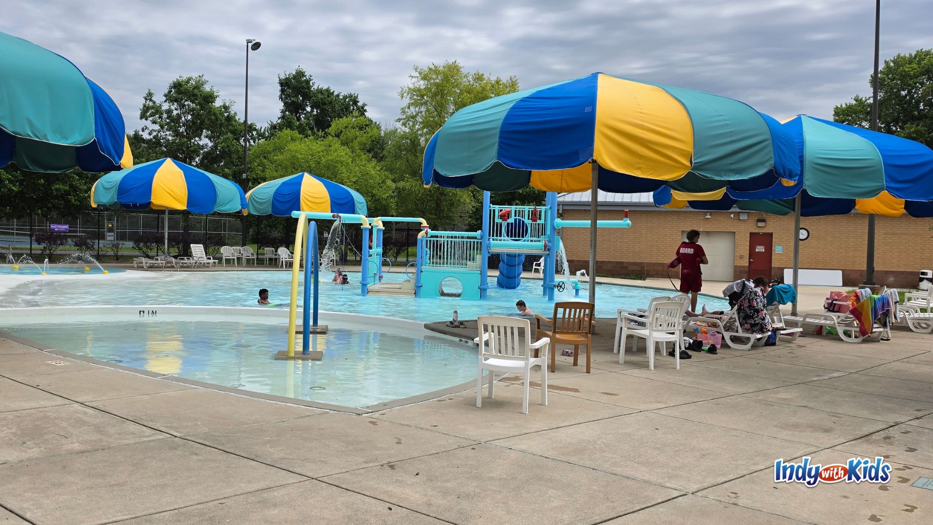 Wide view of the JCC Indianapolis outdoor pool and splash area, with blue, yellow, and teal shade umbrellas, shallow water play structures, lounge chairs, a lifeguard on duty, and families swimming on a cloudy summer day.