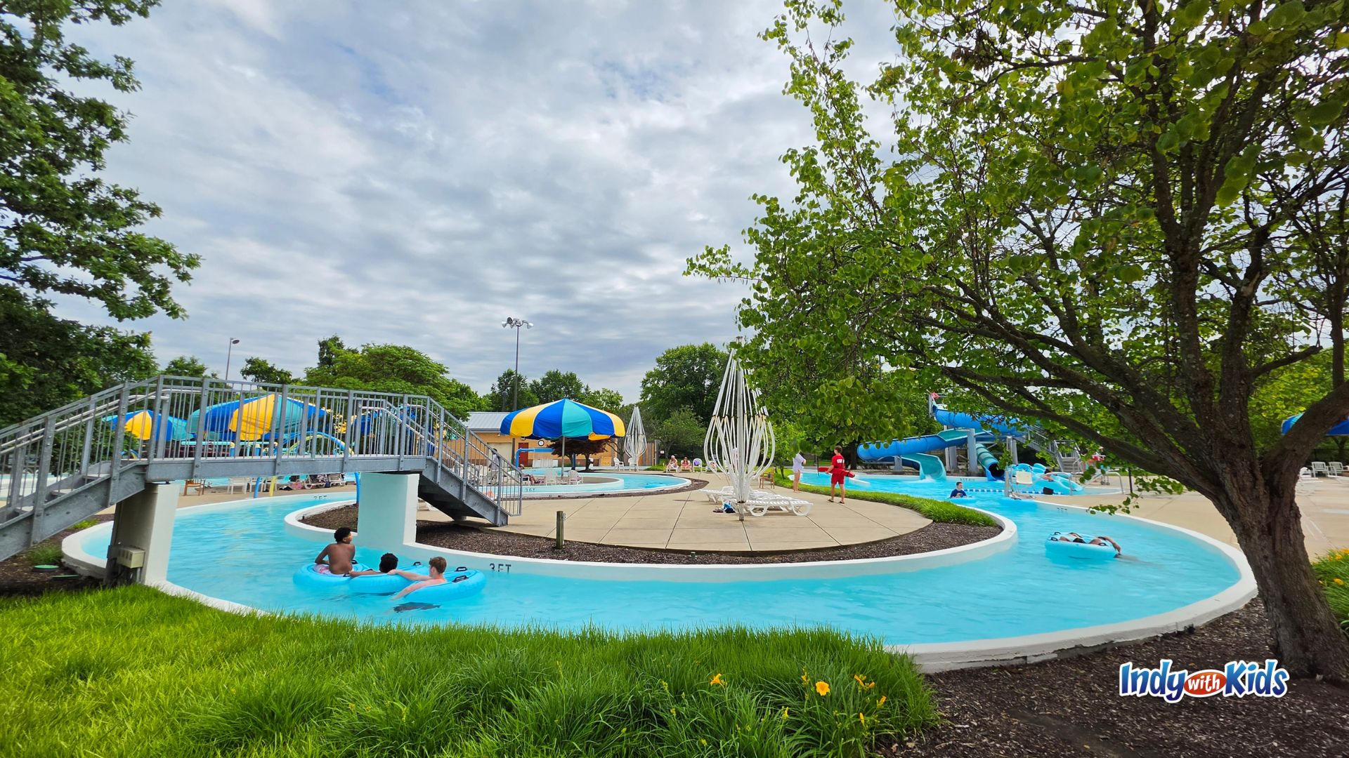 Wide view of the JCC Indianapolis outdoor water park, showing the lazy river, bridge, colorful umbrellas, lounge chairs, water slides, and families floating in tubes on a cloudy summer day.