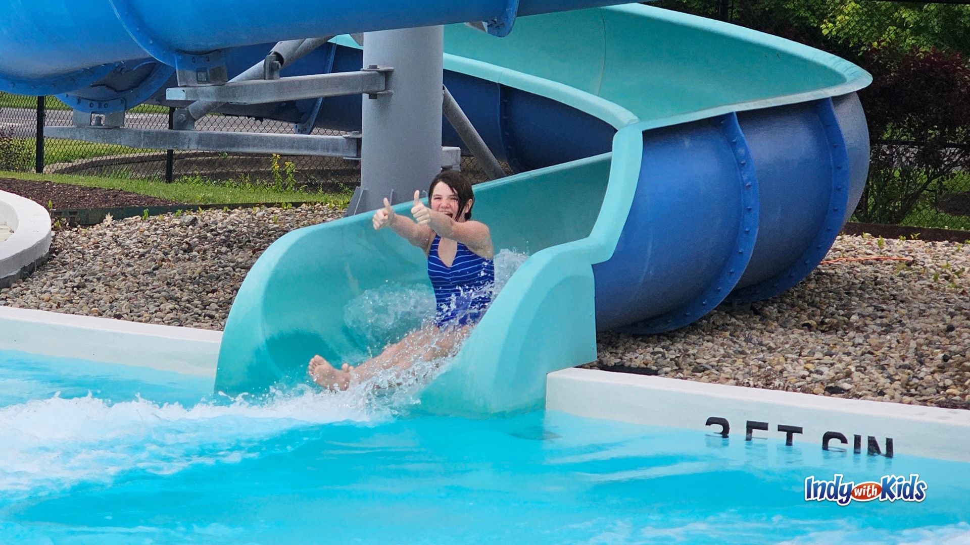 A child smiles and gives two thumbs up while splashing out of a blue and turquoise water slide into the pool at the JCC Indianapolis Water Park.