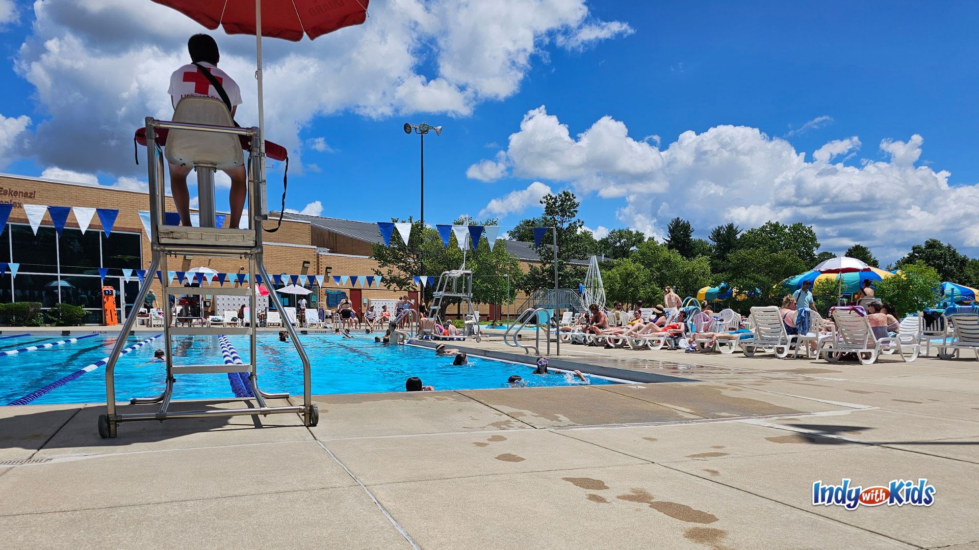 Families swim and relax around the outdoor pool at the JCC Indianapolis, with a lifeguard on duty, lounge chairs, blue-and-white flags, and a bright summer sky overhead.