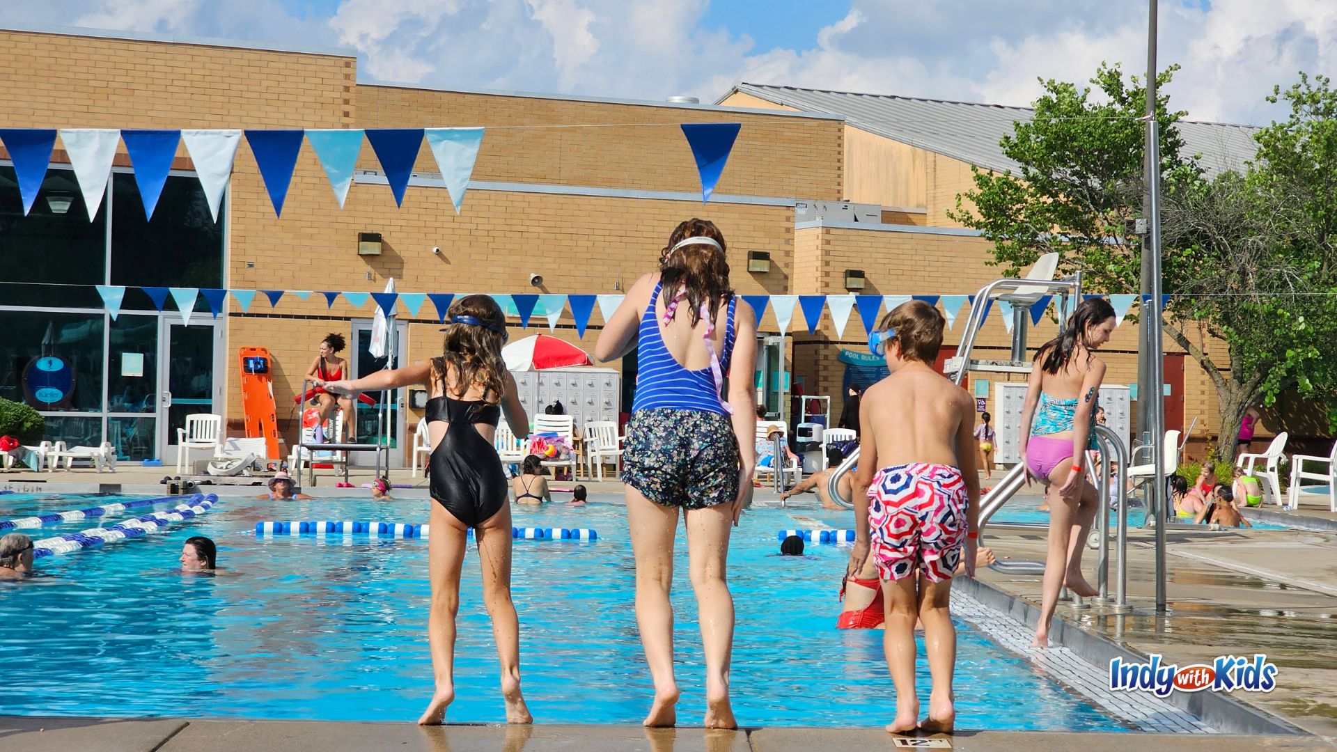 Kids jump in from the edge of the outdoor pool at the JCC Indianapolis, with swimmers in the water, a lifeguard nearby, and blue-and-white pennant flags hanging above the pool deck.