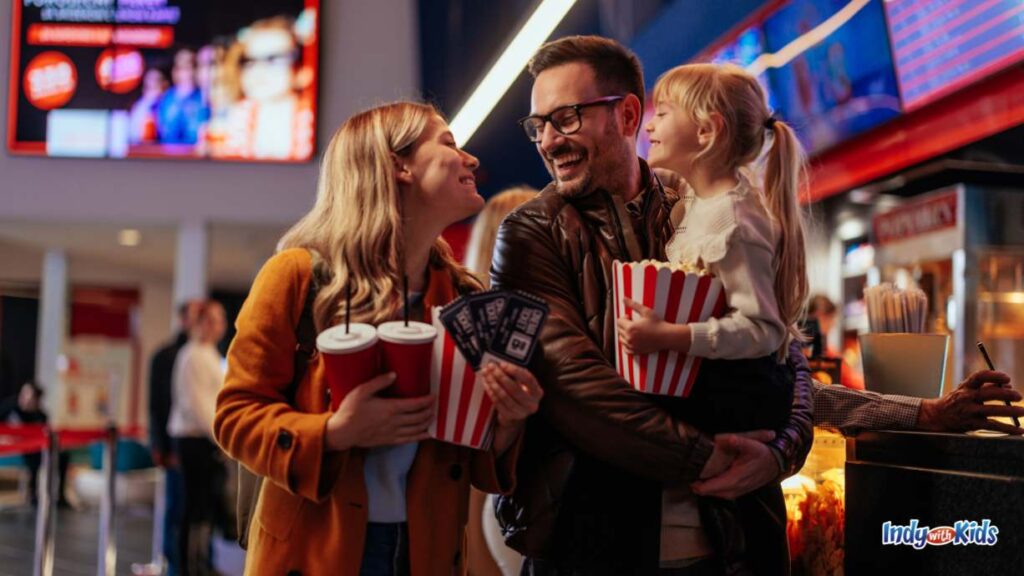 Sensory Friendly Movies: a mother father and little girl being held by the father smile at each other in the movie theater concessions area holding their drinks and popcorm