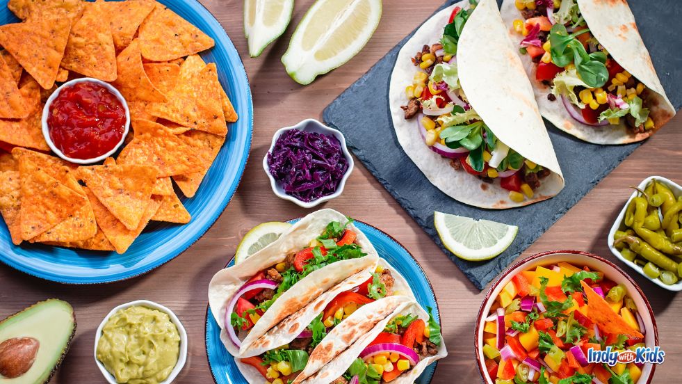 Taco Tuesday: Chips and salsa, avocados, limes, and brightly-colored toppings sit alongside tacos on a wooden table.