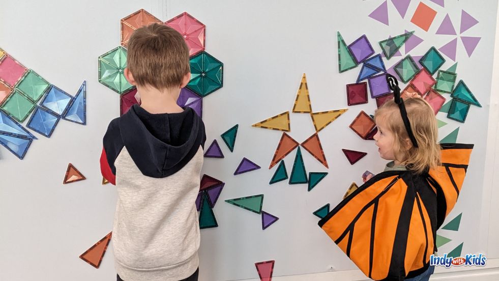 A boy in a sweatshirt and a girl in a butterfly costume play with magnetic shapes on a wall in the Star Studio at Newfields.