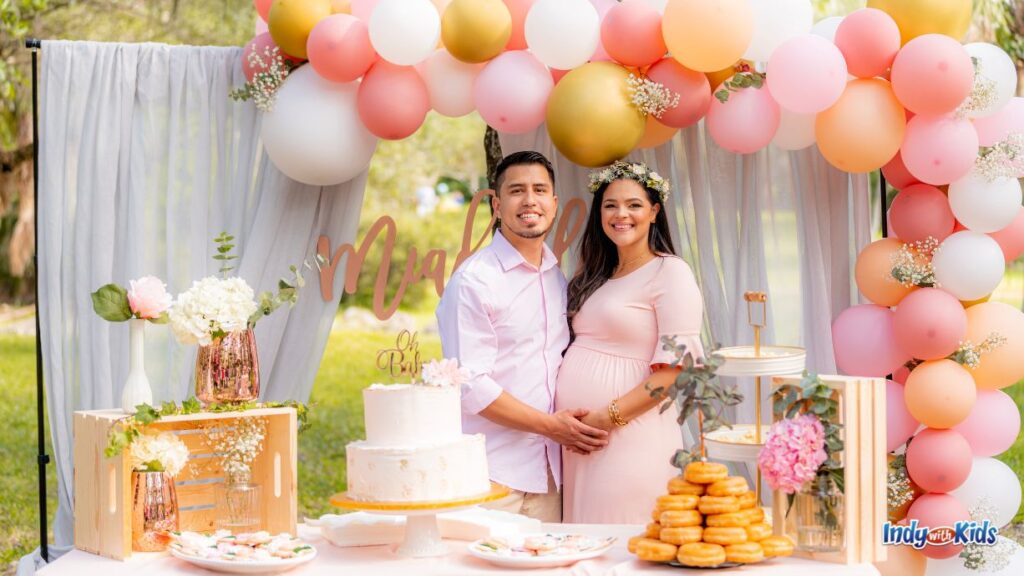 a couple stands together at their outdoor baby shower. there are pink gold and light orange balloons in an arch behind them with a white curtain wall. on the table in front of them are flowers, donuts, cookies, and white cake with an 'oh baby' topper.