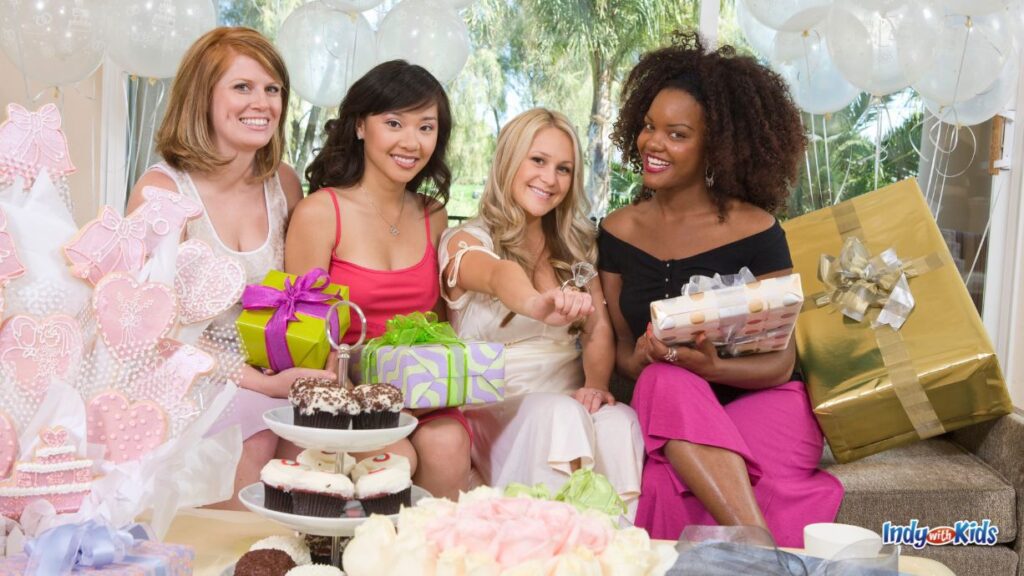 four women sit together at a bridal shower. the bride shows off a giant ring. in front of them are flowers, a three tier dessert tray, gifts, and pink cookies on sticks.