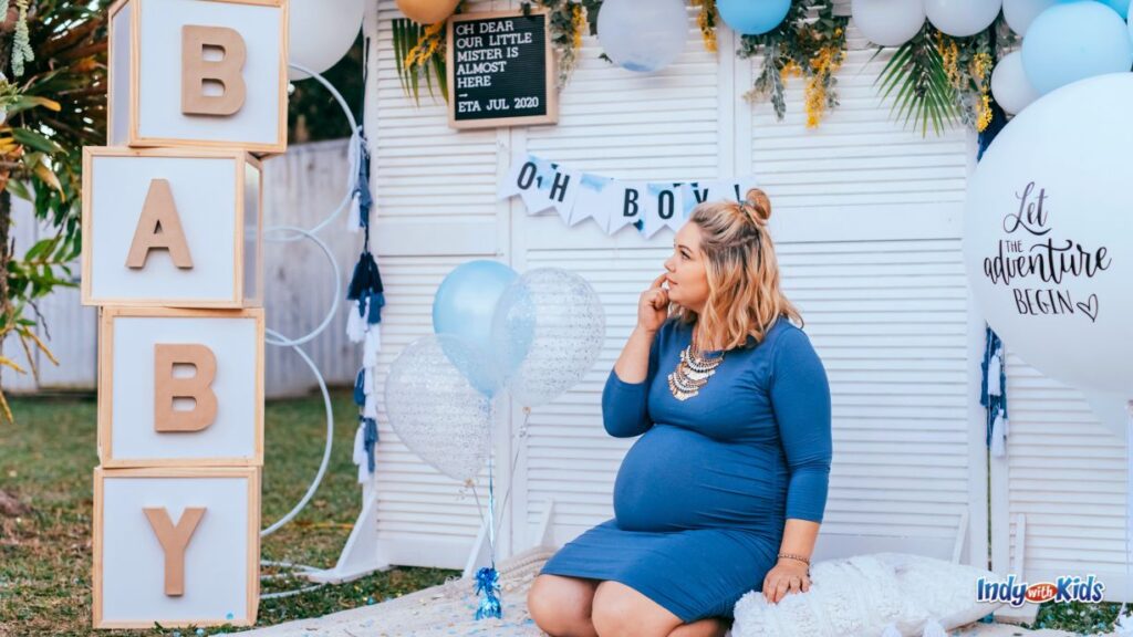 baby shower locations: a mom-to-be in a blue dress sits at a photo spot with giant blocks with the letters b-a-b-y on them. there are balloons and banners with 'oh boy' on them.