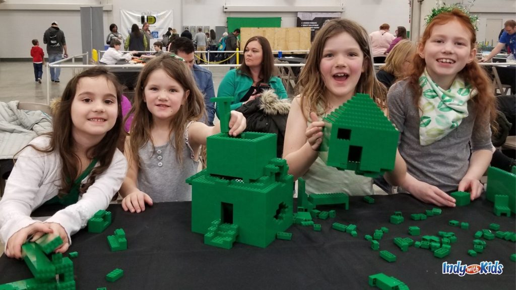 Four kids playing at a table with green LEGO bricks inside an expo hall at Brickworld.