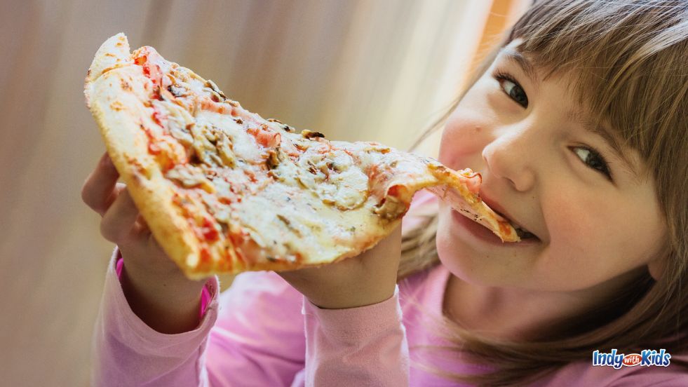 Pi Day Deals Near Me: A girl with brown hair smiles at the camera while taking a bite from a slice of pizza.