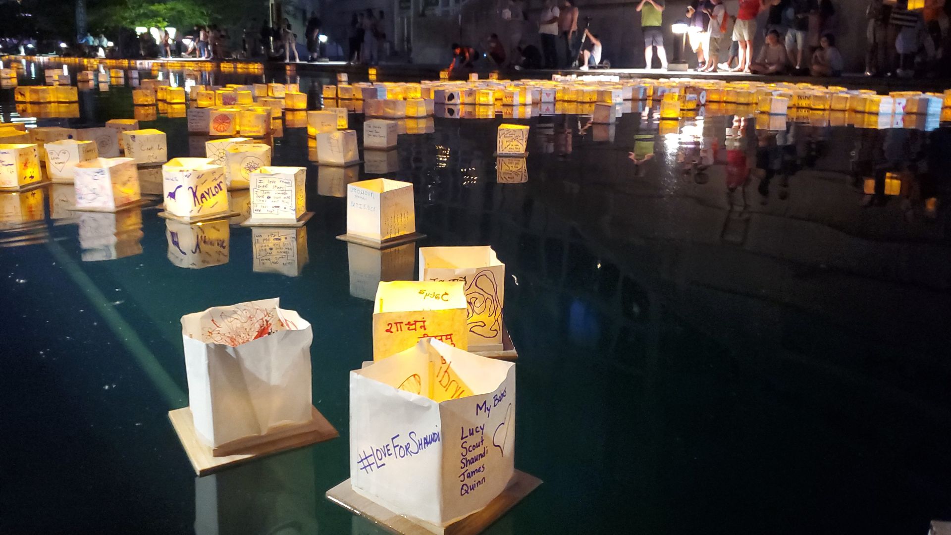 Hundreds of glowing paper lanterns float on the water at the Indianapolis Water Lantern Festival after dark, with visitors gathered along the edge of the canal.