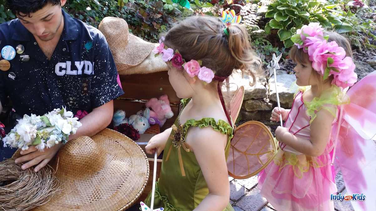 Two girls wearing a flower crowns and costume fairy wings watch as a crafter creates a floral design at Minnetrista's Faeries Sprites and Lights event in Muncie.
