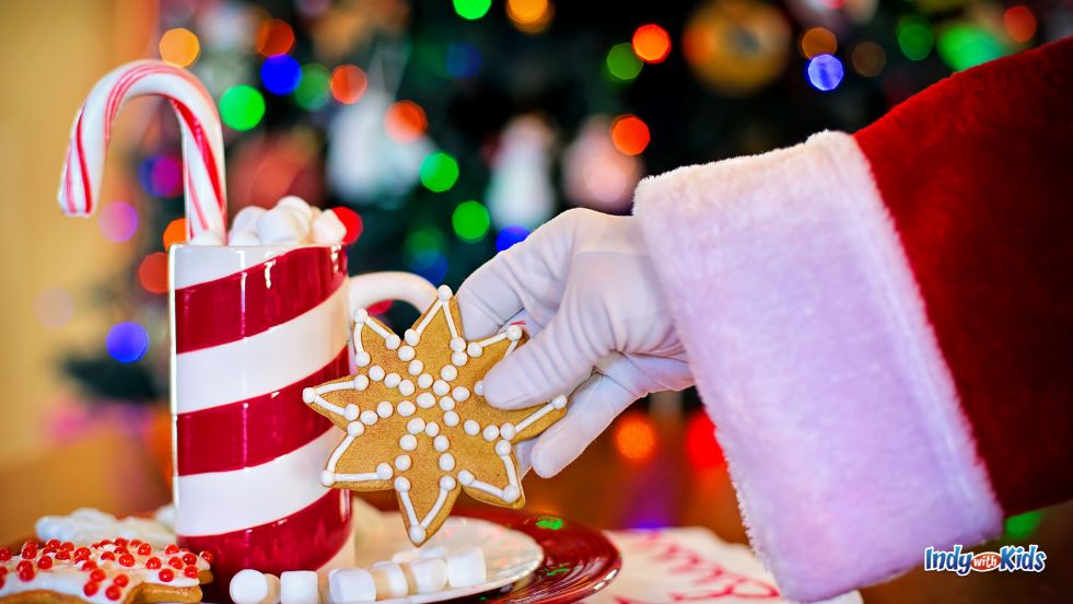 Hendricks County Holiday Events: Santa's white-gloved hand reaches for a star-shaped cookie beside a red striped cocoa cup with a candy cane propped inside.