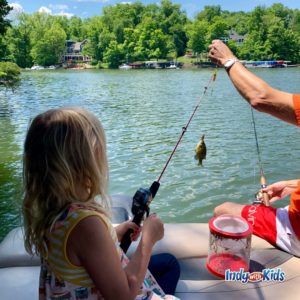 a little girl holds a fishing pole with a little fish hooked on the end. an adult arm is in the picture helping to hold up the fishing pole. they appear to be on a boat on a lake.