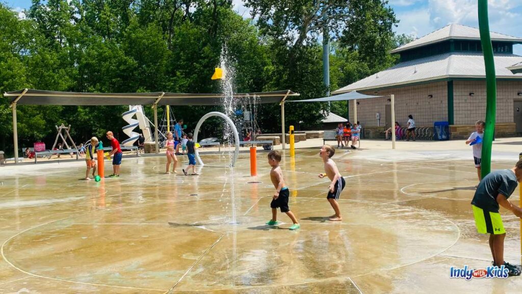 lots of young children are playing in their swim suits on the large splash pad at dillon park. several fountains spray water and one tall stream shoots a yellow cone in the air.
