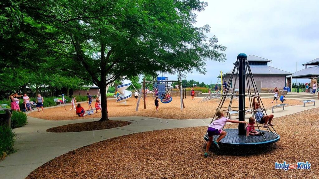 a busy dillon park has parents sitting on benches and children on all of the park equipment like a saucer swing, a pyramid merry go round, and a tall winding slide.