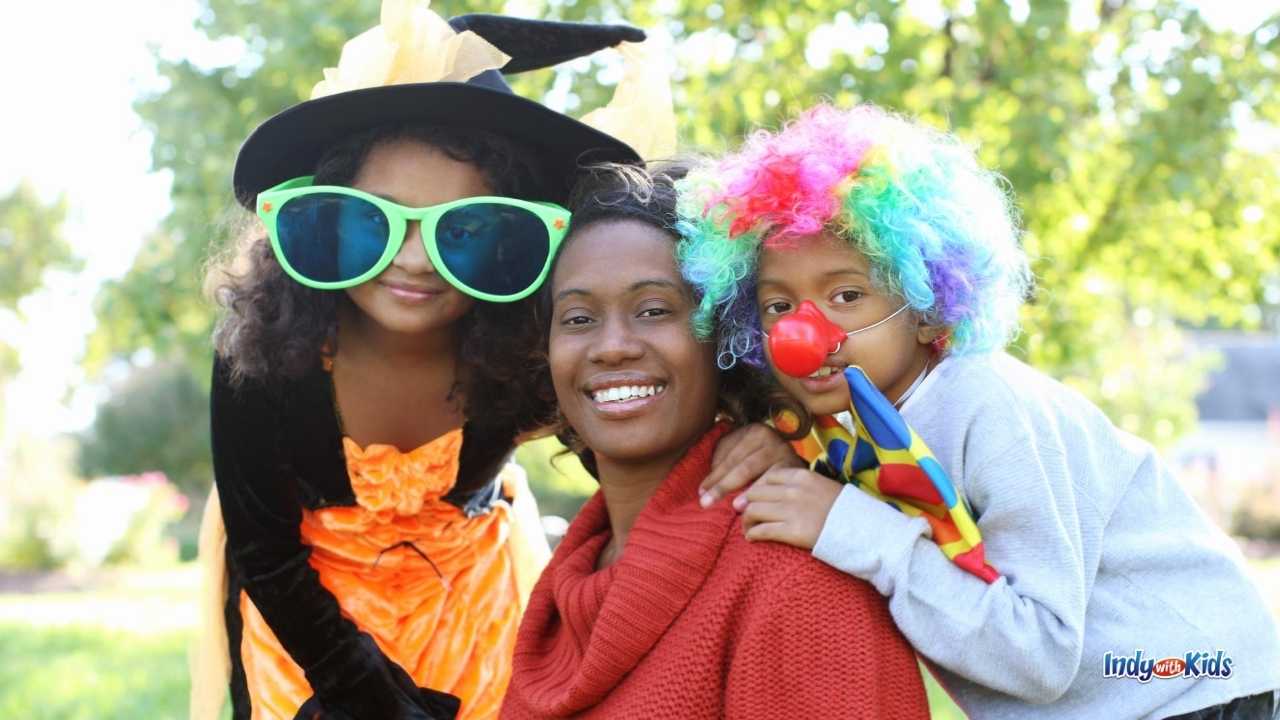 A woman in a sweater with one child in a clown costume and another in a witch's hat and giant sunglasses attend a local Trunk or Treat event.