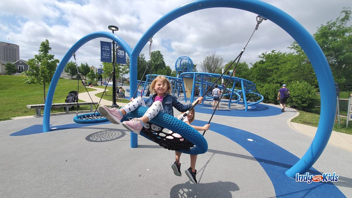 Mud free parks near me: Two girls grin while swinging on a blue circular swing at the Colts Canal Playspace in Downtown Indianapolis.