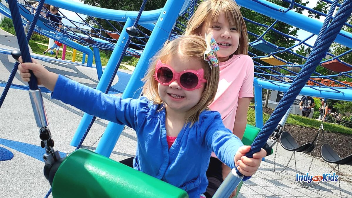 Two children, one wearing pink sunglasses, play on a plastic swing while surrounded by bright blue climbing equipment at the Indianapolis Colts Canal Playspace.