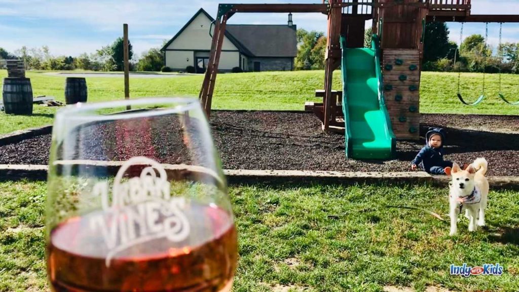 a glass of red wine with the Urban Vines logo is in the forefront of the picture and a dog and a baby are sitting at the playground in the background
