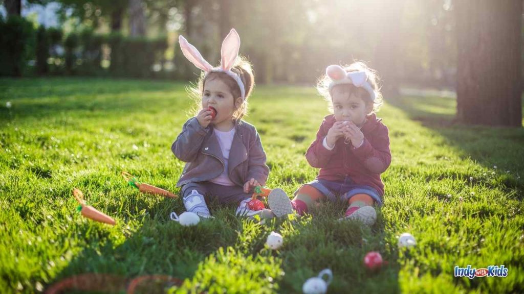 Things to Do in Indianapolis in April: two toddler girls sit in the grass on a lawn with bunny ear headbands eating chocolate from plastic easter eggs