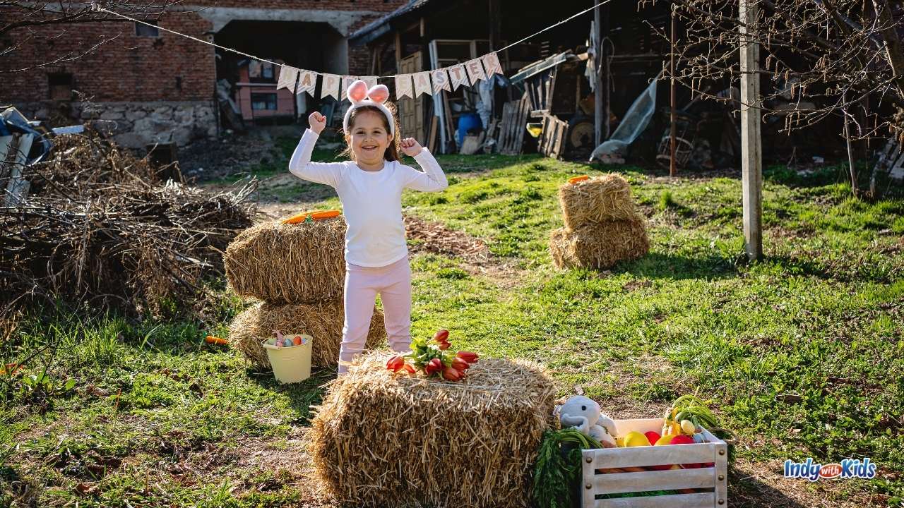 Things to Do on Easter at Home: A girl wearing bunny ears raises her hands in celebration while standing near a full bucket of Easter eggs and several hay bales in a grassy yard.