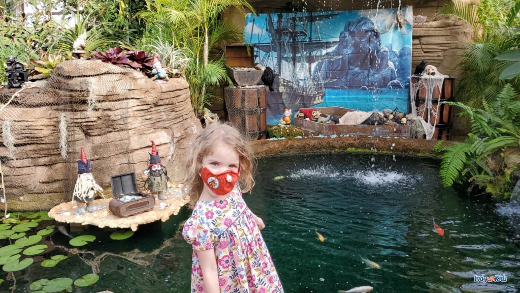 A girl in a pink floral dress poses with the Garfield Park gnomes near a koi pond.
