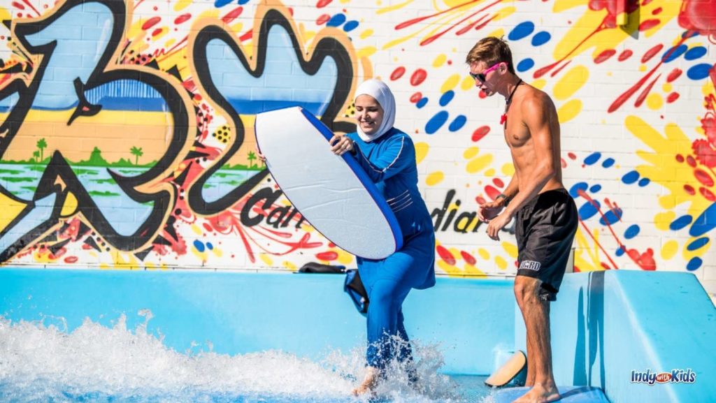 a woman in blue swim shirt and pants and a white head dress prepares to ride the surfing simulator at the Carmel Water Park. she is holding a white body board and a lifeguard with black shorts on is standing next to her ready to help.