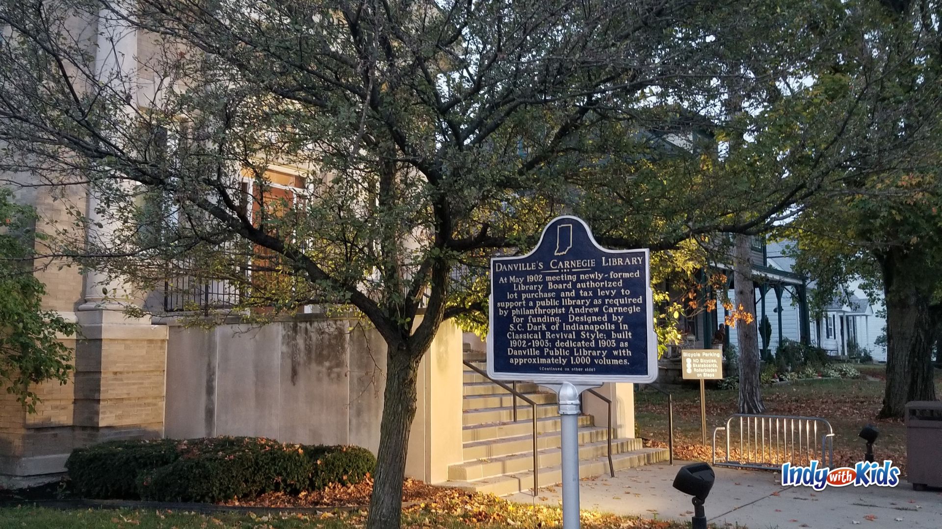 A historical marker for Danville’s Carnegie Library stands outside a stone library building, partially shaded by trees.