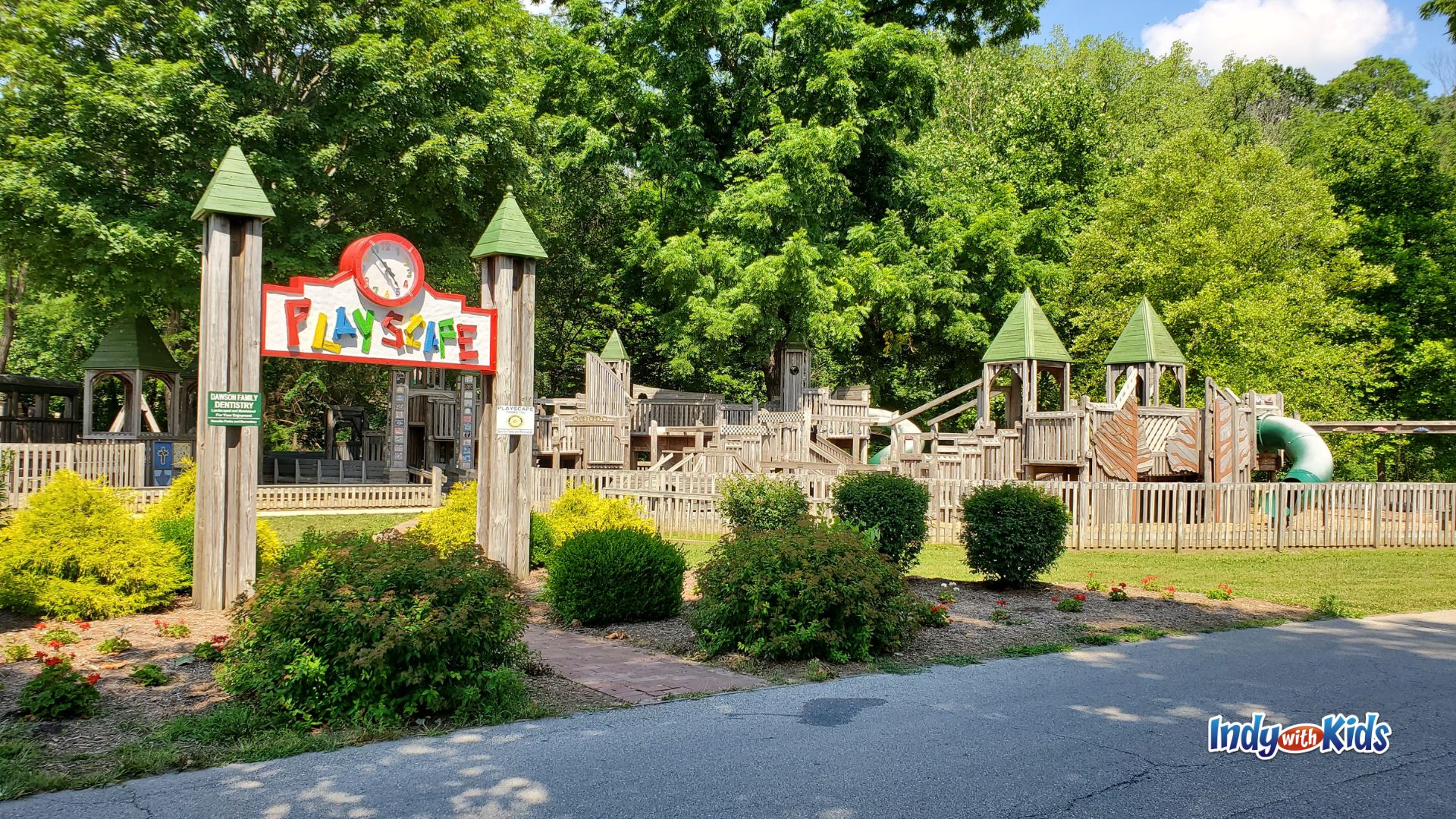 A large wooden playground with castle-like towers, slides, ramps, and a colorful “PlayScape” sign sits surrounded by trees and landscaping at Ellis Park.