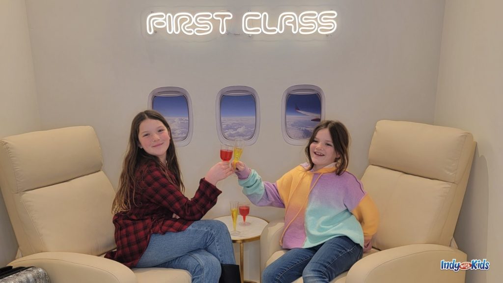 two girls sit in leather reclining chairs are cheersing with juice flutes in front of an airplane window backdrop and a lit up white sign that says "first class"