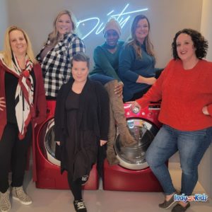 six women pose in front of and on top of red washing and drying machines. on the wall in white cursive handwriting is the word "dirty"