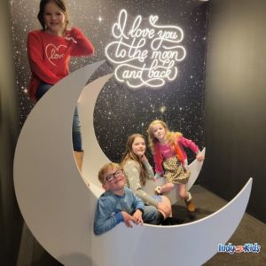 3 children sit in and one sits atop a moon shaped bench. the backdrop wall is black with white stars and the phrase "i love you to the moon and back" lit up in white 