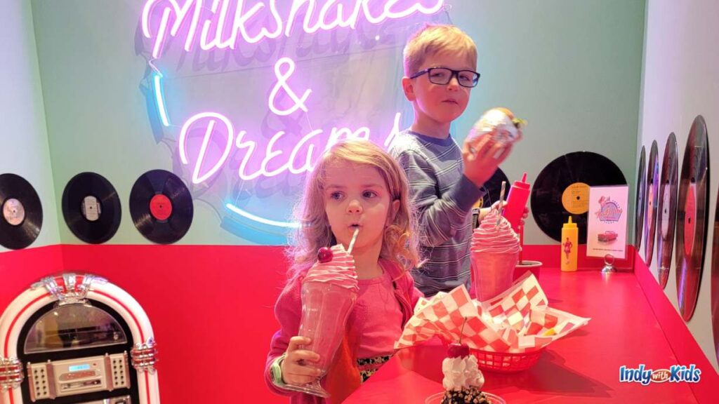 a girl and a boy sit at a red counter in a diner setting, with a jukebox on the ground next to them, pretend-sipping on a milkshake and eating a burger. there are records on the wall and a sign that says "milkshakes & dreams"