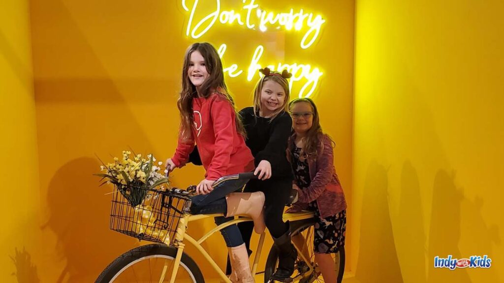 three elementary aged girls sit atop an adult sized bicycle with a basket and yellow flowers inside in front of a yellow wall and a lit up sign that says "don't worry be happy"
