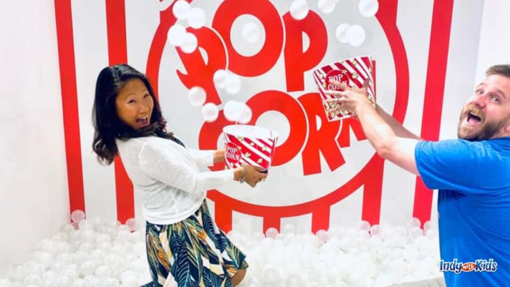 a couple stand in a pit of white ball pit balls at selfie wrld indy holding popcorn tubs and throw the white balls up in the air in front of a red and white popcorn bag design