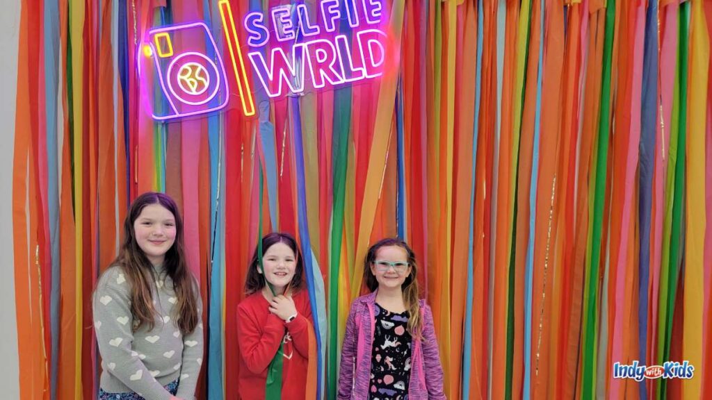 three girls stand in front of a colorful wall of rainbow colored ribbons hanging down and a lit up sign that says selfie wrld with a camera image