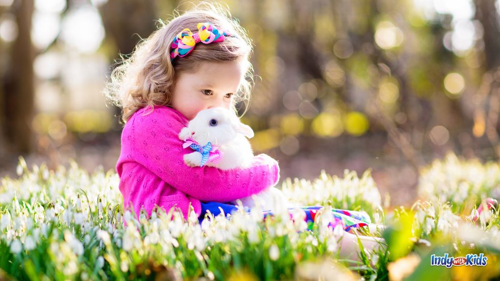 Easter bunny pictures near me: A small girl in a pink cardigan sits in the grass and kisses the head of a white bunny wearing a bow around his neck.