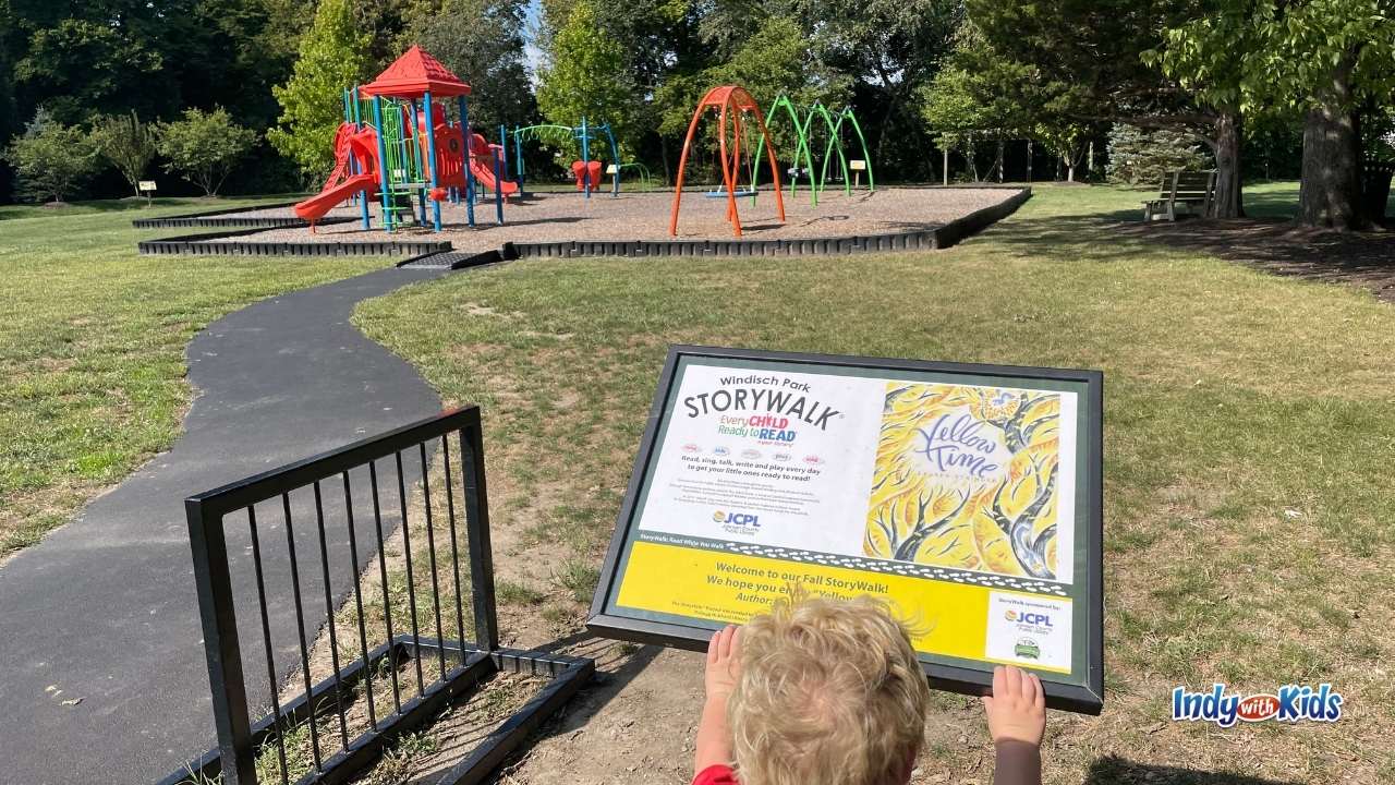 Story Walk Near Me: A child reads a page from a storybook displayed on a sign in front of a playground at a local park.